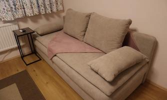 A beige sofa with large cushions and a blanket in pink. Next to the sofa stands a small table.