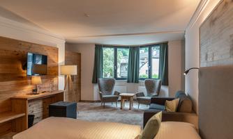 A modern hotel room with a comfortable bed, a cozy seating area, and large windows. The atmosphere is inviting and bright, with wood and fabric elements.
