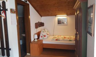 A cozy room with a single bed, a wooden ceiling, and a nightstand. A picture hangs on the wall and there is access to a bathroom.