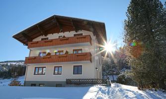 A beautiful house in the snow with wooden balconies. The sun is shining brightly and there are tall trees nearby.