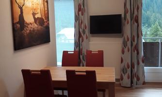 A cozy dining area with a wooden table and four red chairs. On the wall hangs a picture of deer, and there are windows with a view of nature.
