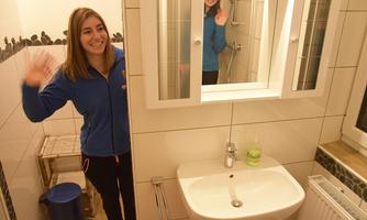 A young woman is smiling in a bathroom and waving. Her reflection is visible in the mirror.