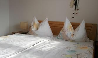 A cozy bedroom with a large double bed and decorative pillows. In the background, there is a picture frame with photo frames and decorations.