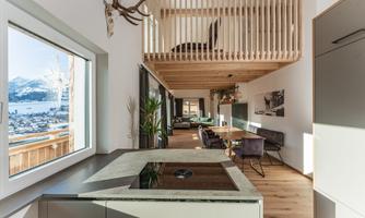 A modern living room with wood decor and large windows that offer a view of a mountain landscape. There is an open living area with a dining table and a kitchen.