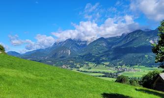 A picturesque landscape with green meadows and majestic mountains in the background. The sky is clear and sunny, with a few clouds.