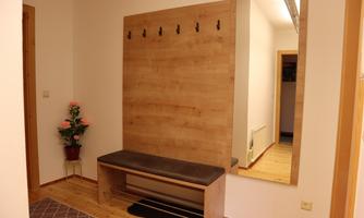 A bright entrance area with a wooden bench and wall hooks. In the background, there is a mirror and a plant stands in the corner.