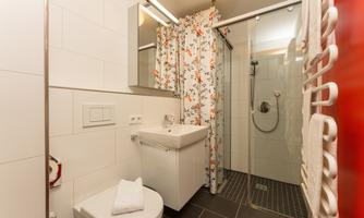 A modern bathroom with a shower, a sink, and a toilet. The wall covering is light and there is a decorative curtain with a floral pattern.