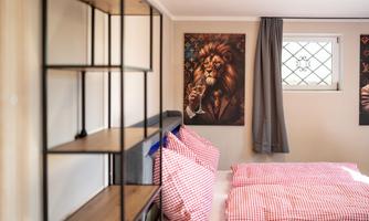 A modern bedroom with a cozy bed and red checkered pillows. Pictures hang on the wall, including a portrait of a lion in a suit.