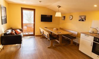 A cozy kitchen-living room with a dining table, a couch, and a balcony. The walls are painted in warm yellow tones and the atmosphere is inviting.