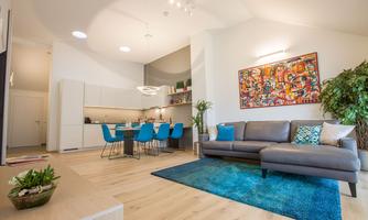 A modern living room with a comfortable couch and a large rug. In the background, you can see a dining area with blue chairs and a kitchenette.