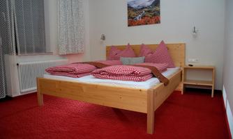 A cozy bedroom with a wooden bed and checkered pillows. The floor is red and a picture is hanging on the wall.