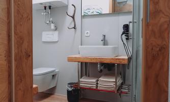A modern bathroom with a wooden sink cabinet and a white wash basin. Stacked towels are resting on the shelf and a toilet area is visible.