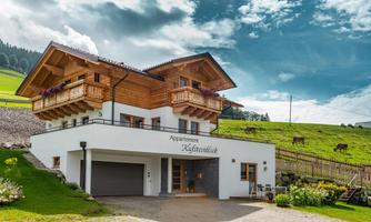 A modern apartment building with traditional wooden design in a rural setting. The buildings are surrounded by meadows and a beautiful sky.