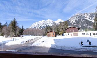 A wintry landscape with snow-covered mountains and wooden chalets. The sky is clear and blue, highlighting the tranquil atmosphere.