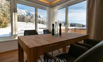 A modern dining area with a wooden table and stylish chairs. Through large windows, there is a view of a snow-covered landscape and mountains.