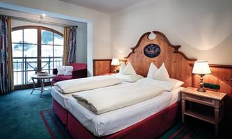 A cozy hotel room with two beds and traditional wooden furniture. Large windows provide a view outside and ensure plenty of daylight.