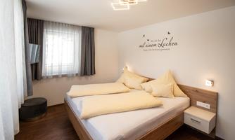 A modern bedroom with two single beds, large windows, and simple furniture. The walls are painted in light colors and feature a motivating quote.