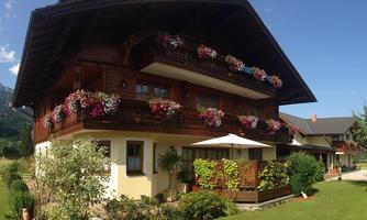A rustic wooden house with balconies, adorned with colorful flowers. In the foreground, there is a well-maintained garden with greenery and a sun umbrella.