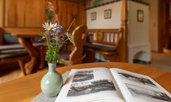 A cozy table with a small bouquet of flowers in a vase and an open book. In the background, wooden paneling and a seating area can be seen.