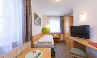 A simple room with a single bed, a table, and a television. There is a window with curtains and a bright, cheerful atmosphere.