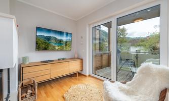 A modern living room with a wooden floor and a TV on the wall. Through the large glass door, one reaches a balcony with a view of nature.
