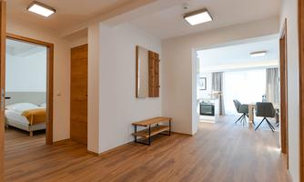 A modern hallway with wooden flooring and bright walls. In the background, a living room with a window and dining area is visible.