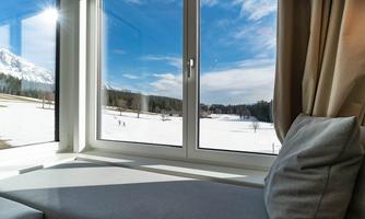 A cozy view through a large window onto a snowy landscape. In the background, trees and mountain peaks are visible under a blue sky.