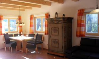 A cozy living space with a dining table, chairs, and a rustic cupboard. Bright windows and orange curtains create an inviting atmosphere.