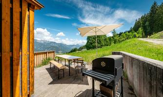 A terrace with a grill, tables and chairs, surrounded by a beautiful mountain landscape. The sky is clear and there is sunshine.