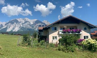 A beautiful house with a balcony and colorful flowers in the foreground. In the background, majestic mountains can be seen under a clear blue sky.
