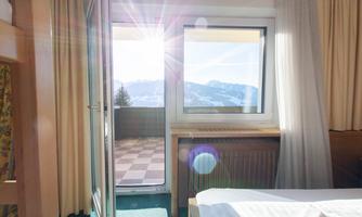 A bright room with a window and a balcony that offers a view of the mountains. The sun rays shine through the window, creating a cozy atmosphere.