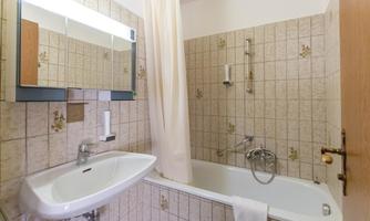 A bathroom with a bathtub, a sink, and a mirror. The walls are tiled and the atmosphere is simple and functional.