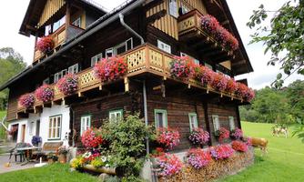 A rustic wooden house with blooming balcony boxes full of flowers. The surroundings are green and rural, with a well-kept garden.