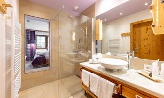 A modern bathroom with a large glass shower and an elegant washbasin. In the background, a cozy bedroom with wooden details can be seen.