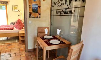 A cozy dining area with a table and chairs. In the background, there is a bedroom with a bed and a decorative glass front on the wall.