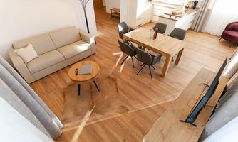 A modern living room with a beige sofa, a wooden table, and elegant chairs. The floor is made of wood and the room is bright and welcoming.