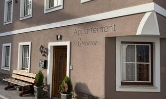 An apartment building with the inscription "Appartement Romana". In front of the entrance, there are potted plants and a wooden bench.