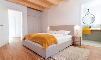 A modern bedroom with a comfortable bed and a warm yellow tone for the bedding.  
Large windows and wooden ceilings create an inviting atmosphere.