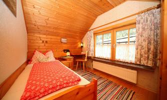 A cozy bedroom with wooden paneling and a single bed. In front of the window are pretty curtains and a small desk.