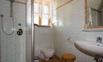 A bright bathroom view with a shower, a toilet area, and a sink. The room has light tiles and large windows with curtains.