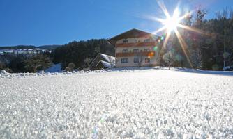 A snow-covered landscape with bright sunlight. In the background, a cozy house and forests can be seen.