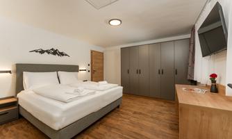 A modern bedroom with a large bed and light wooden floor. The room features a television, a wardrobe, and cozy decoration.