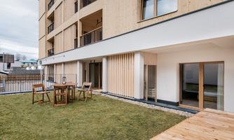 A modern terrace with a small lawn area and wooden furniture. In the background, a multi-story building can be seen.