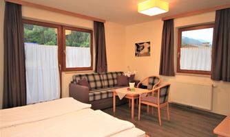 A cozy room with two beds, a sofa, and a table in a bright space. Large windows provide plenty of daylight and a view of the greenery.