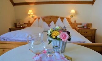 A cozy bedroom with a large bed and warm wooden decor. On the table, there is a flower vase and a carafe with glasses.