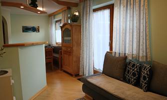 A cozy living space with a sofa and a wooden cabinet. Bright windows and decorative curtains create an inviting atmosphere.