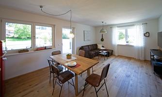 A cozy living-dining area with wooden floor, a table, and chairs. Large windows provide plenty of daylight and a view of the greenery.