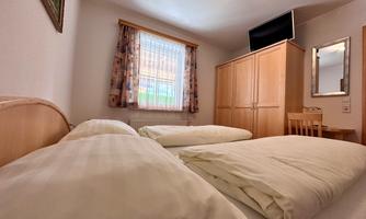 A simple bedroom with two beds and a window. There is a wardrobe, a table, and a television on the wall.