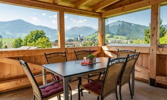 A cozy dining area on a covered terrace overlooking green mountains and a clear blue sky. The table is decorated with a plant and surrounded by chairs.