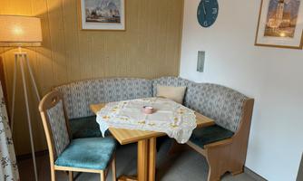 A cozy dining area with a wooden bench and a table. Pictures and a clock hang on the wall, while a lamp provides soft light.